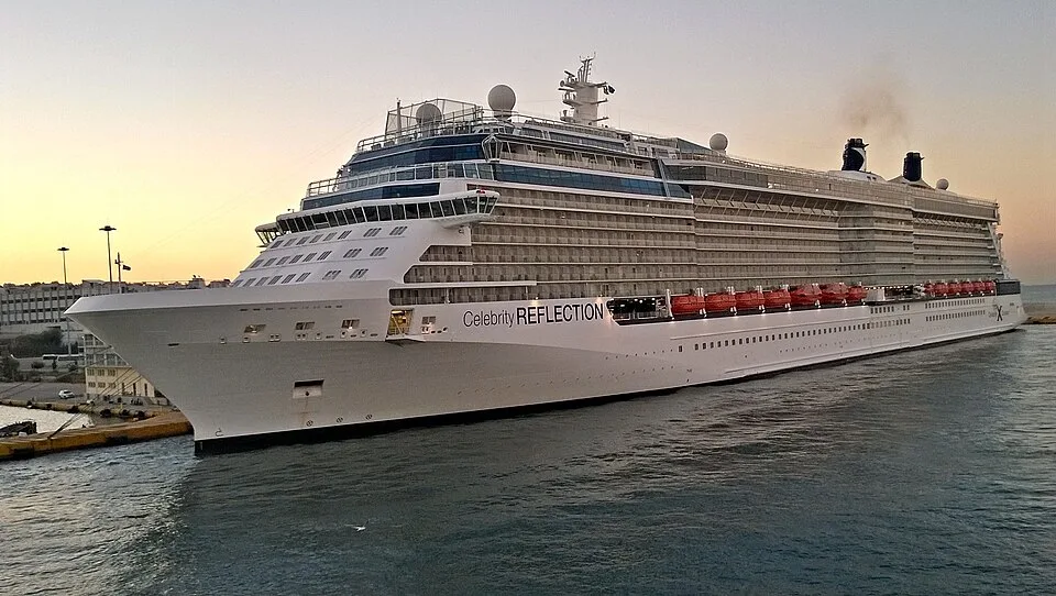 The massive white cruise ship Celebrity Reflection is docked in a harbor during sunrise or sunset. The vessel features multiple decks lined with glass balconies, orange lifeboats along the side, and two dark smokestacks near the rear emitting faint exhaust under a pale sky.