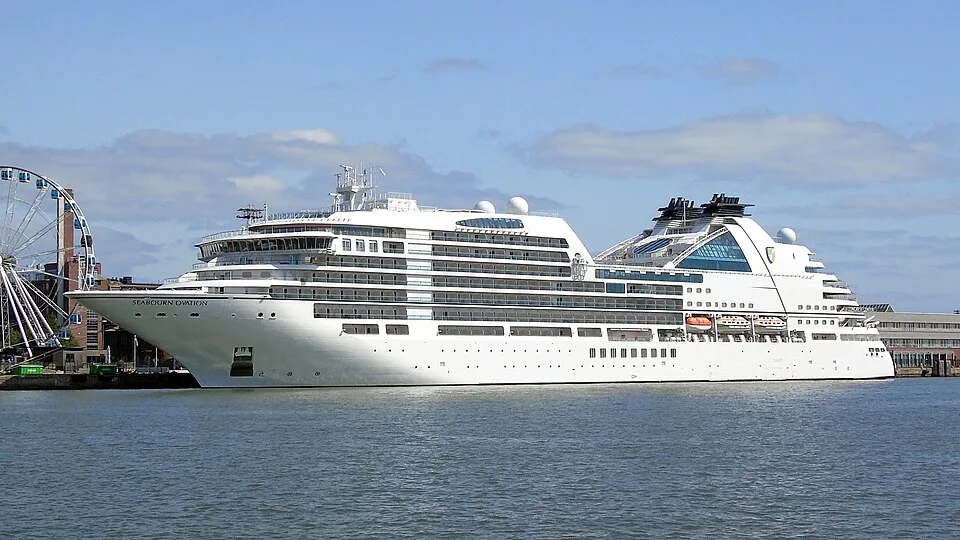 A sleek white cruise ship named "Seabourn Ovation" docked along a waterfront under a partly cloudy blue sky. The vessel features modern tiered decks, private balconies, and orange lifeboats, with a large Ferris wheel partially visible on the far left.