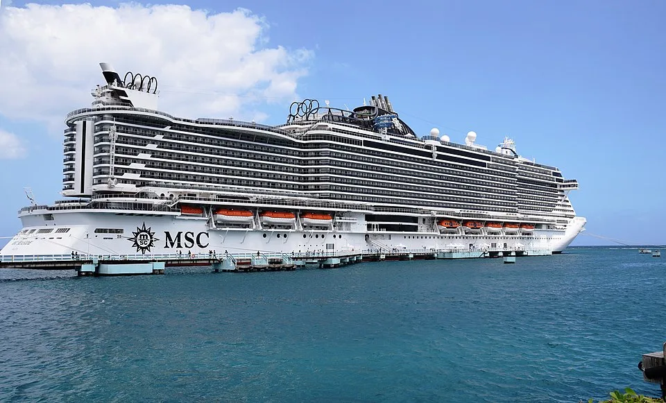 A large MSC cruise ship docked at a pier on a bright, sunny day. The massive white and black ship features multiple decks with balconies and bright orange lifeboats. It sits in clear blue ocean water under a sky with light, fluffy clouds.