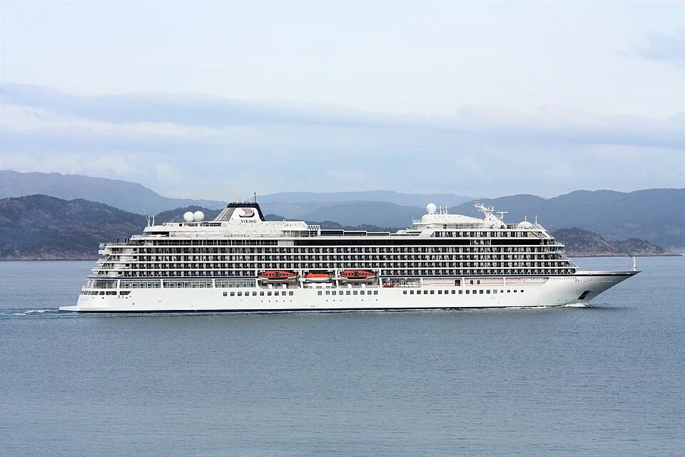 A massive white Viking cruise ship sails on calm, open water past a rugged, mountainous coastline under an overcast sky. The long vessel features multiple tiers of passenger balconies, a row of orange lifeboats along the middle deck, and a dark funnel bearing the Viking logo.