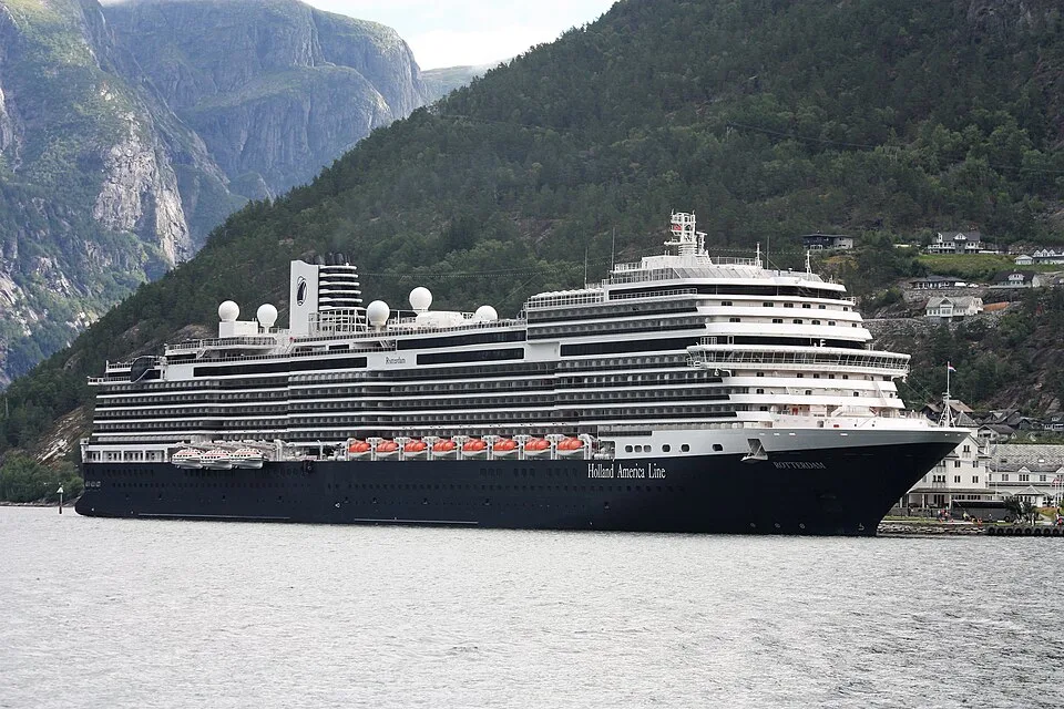 The large Holland America Line cruise ship "ROTTERDAM" sits in a fjord surrounded by water. The ship features a dark hull, tiered white upper decks, orange lifeboats, and white satellite domes. Steep green, rocky mountains and scattered houses line the shore in the background.