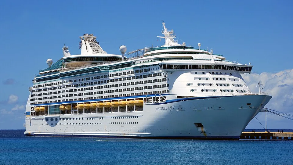 A large white Royal Caribbean cruise ship, named "Adventure of the Seas," docked in calm blue water under a clear blue sky. The multi-level ship features rows of balconies and yellow lifeboats along its side.