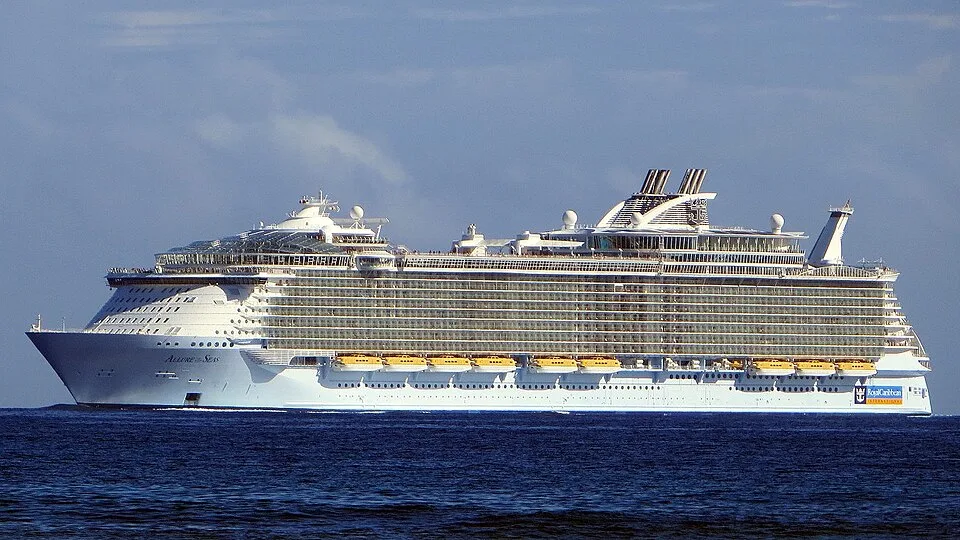The massive Royal Caribbean cruise ship Allure of the Seas sails on deep blue water under a hazy blue sky. The multi-deck white vessel features its name near the bow, rows of yellow lifeboats along the side, and prominent split funnel structures on top.