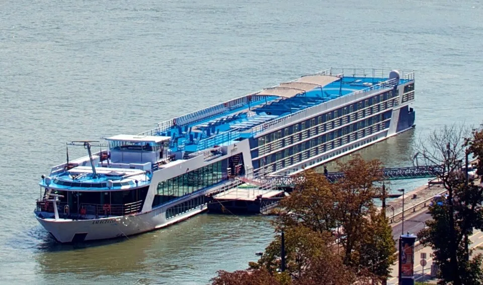 An aerial view shows a long, white and blue river cruise ship docked along a riverbank pier. The multi-deck vessel features a bright blue sun deck with lounge chairs, glass-walled lower common spaces, and multiple tiers of passenger cabins lined with windows under bright sunlight.