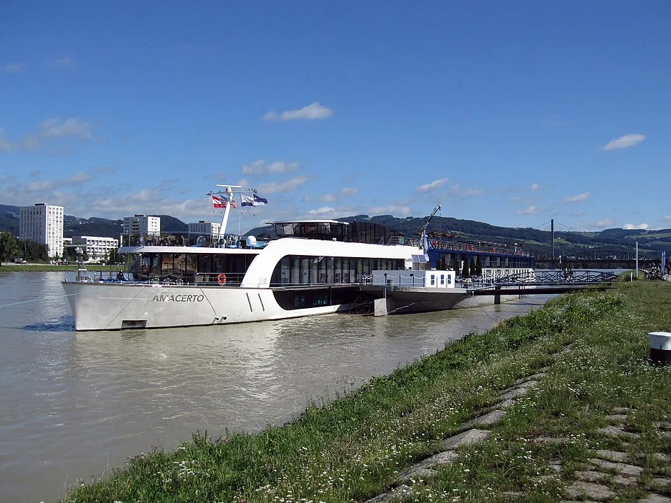 A white river cruise ship named "AmaCerto" docked along a grassy riverbank. The modern, low-profile vessel features glass walls, multiple flags flying from its mast, and a gangway connecting it to the shore, with buildings and hills under a bright blue sky in the background.