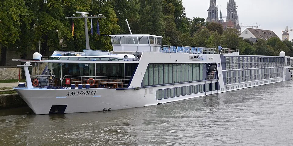 The river cruise ship AmaDolce is docked along a tree-lined bank on calm water. The white and blue vessel features large glass windows on its lower decks and rows of passenger cabins toward the back. Historic church spires rise above the lush green trees in the distant background.