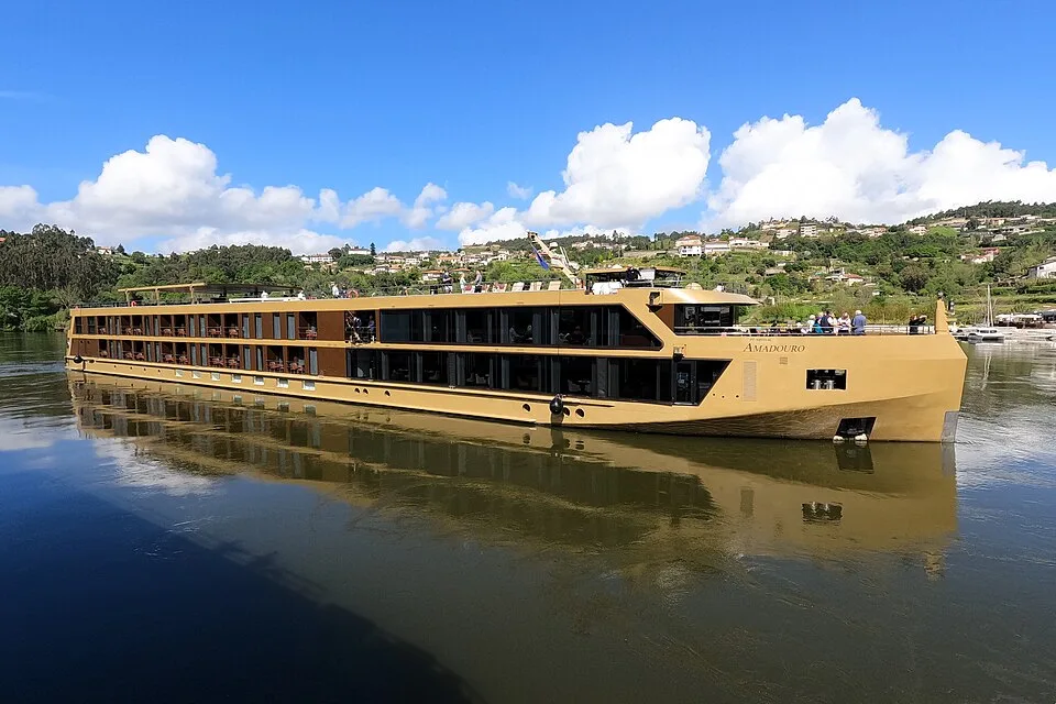 A golden-tan river cruise ship named "AmaDouro" sailing on a calm river. The low-profile vessel features glass panels and passenger cabins, casting a clear reflection on the water with a green, house-dotted hillside under a bright blue sky with white clouds in the back.