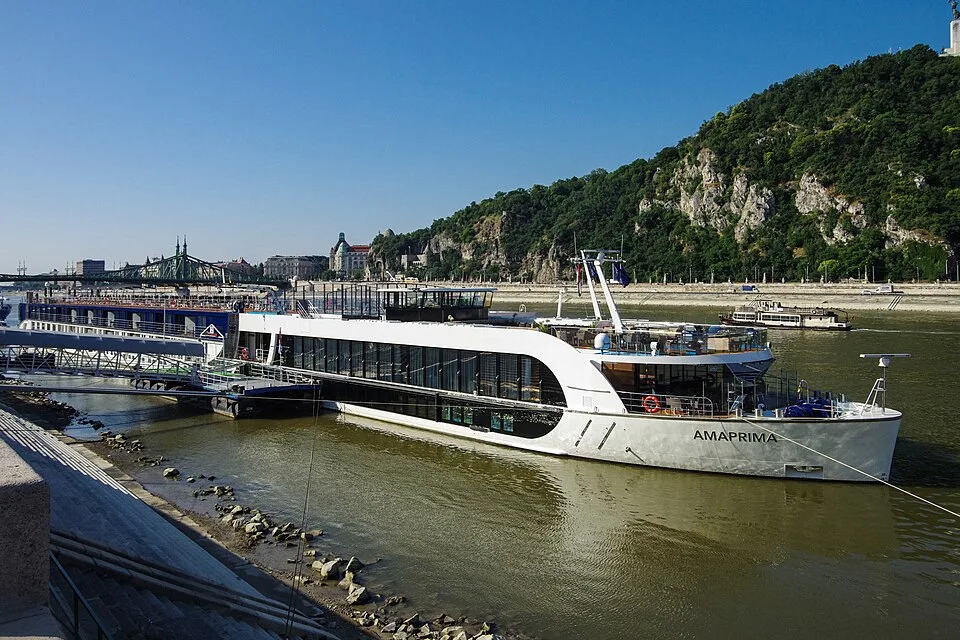 A luxury white river cruise ship named AMAPRIMA is docked along a riverbank next to a green hill. In the background, a green bridge stretches across the water under a clear blue sky.
