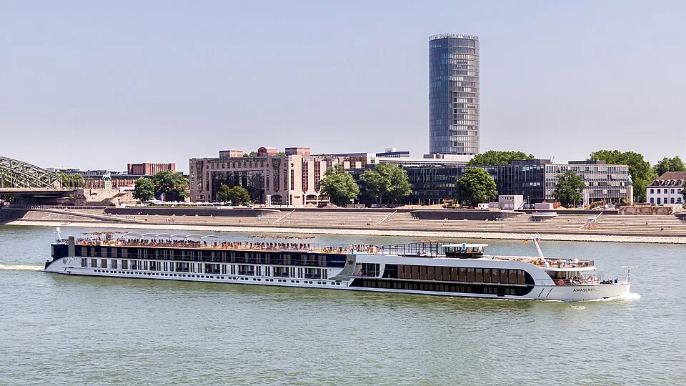 An AmaSerena cruise ship sails along a wide river, with a city skyline in the background featuring a tall cylindrical glass tower. The white vessel has multiple decks, passenger balconies, and a sun deck. The foreground shows calm water under a clear, bright sky.