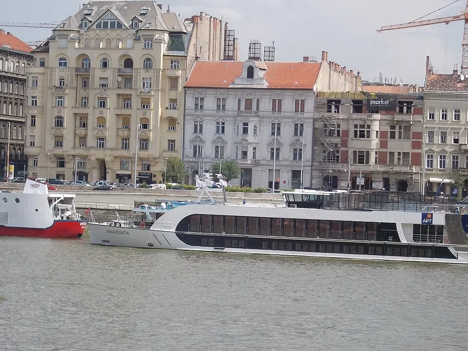 The AmaVenita river cruise ship is docked on a wide river in front of historic European buildings with orange-tiled roofs. A smaller red and white boat is visible nearby to the left, and a construction crane rises into the sky in the background.