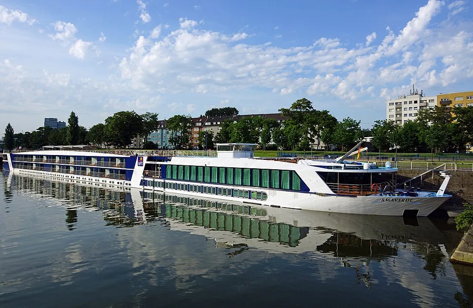 Side view of the white and blue AmaVerde cruise ship docked on a calm river. Green trees, city buildings, and a blue sky with fluffy clouds reflect clearly on the water's surface, with the ship's name visible on the bow.