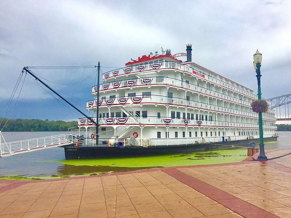 A large, white multi-deck riverboat with red trim and patriotic bunting is docked along a paved walkway. The ship features ornate Victorian-style railings, multiple levels of balconies, and a black hull resting in green-tinted river water under a cloudy sky.