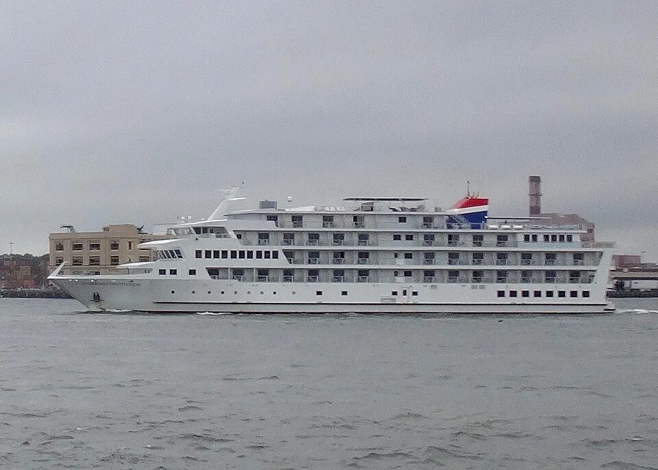 A large white cruise ship sails across gray water under an overcast sky. The multi-deck vessel features rows of small balconies and a distinct red, white, and blue smokestack near the rear, with buildings visible on the distant shoreline.