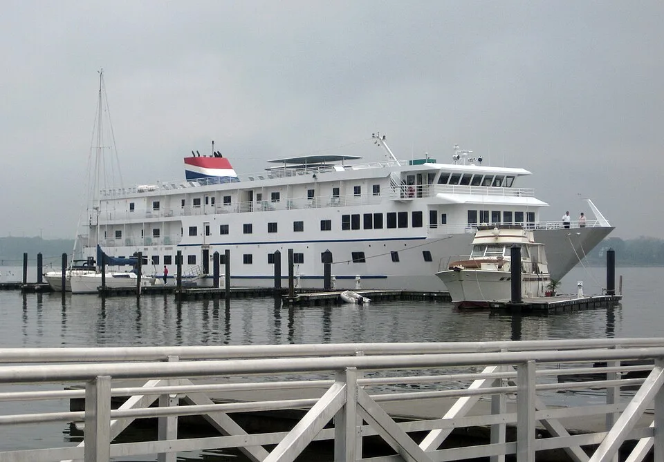 A large, white multi-deck cruise ship with a red, white, and blue smokestack is docked at a pier under an overcast sky. A smaller motor yacht and a sailboat are moored alongside it, with a metal railing visible in the foreground.