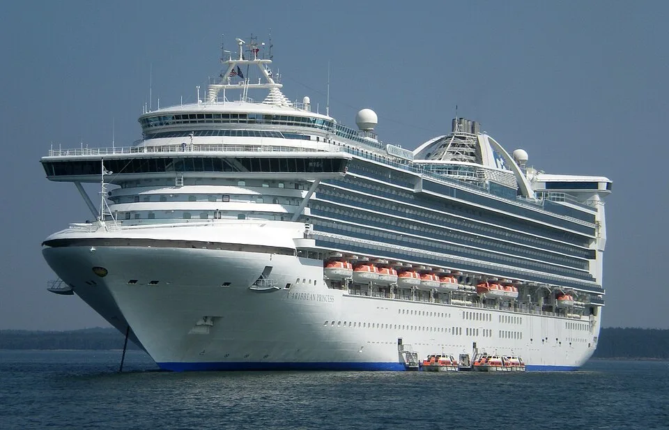 The cruise ship Caribbean Princess sits anchored in calm water. The massive white vessel features a wide glass bridge, multi-level windowed decks, and a row of orange lifeboats along the side. Two small tender boats are docked at its base.