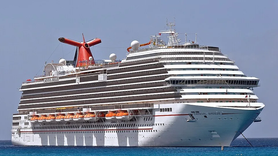 A large white cruise ship named "Carnival Breeze" sailing on bright blue water. The massive vessel features many tiered decks with rows of balconies, orange lifeboats along the side, and a distinctive red and blue whale-tail smokestack on top under a clear sky.