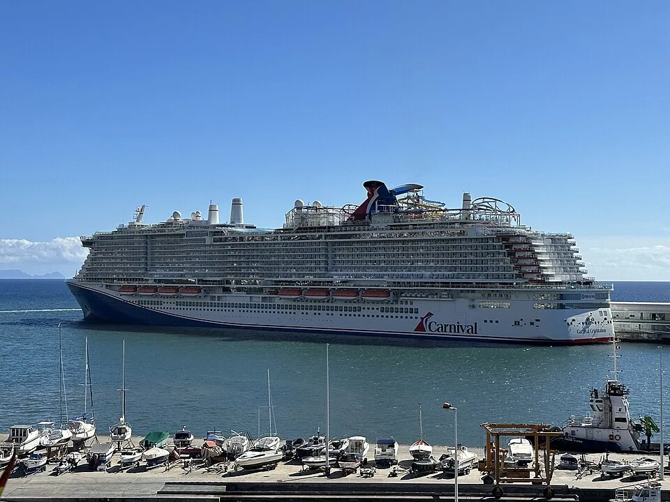 A massive white and blue Carnival cruise ship is docked in a harbor under a clear blue sky. Smaller boats and a tugboat are lined up at a concrete marina in the foreground, with distant land visible on the ocean horizon.