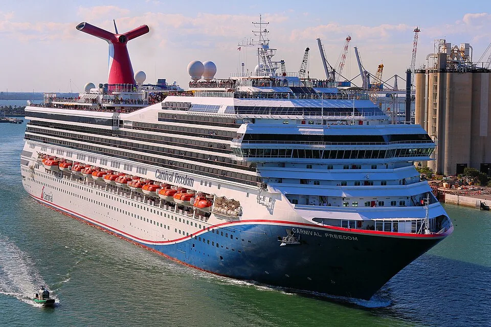 A massive Carnival Freedom cruise ship with a dark blue, white, and red hull navigating a harbor channel. The ship features tiered cabin decks, orange lifeboats, and a signature red whale-tail smokestack, with a small pilot boat nearby and an industrial port in the background.