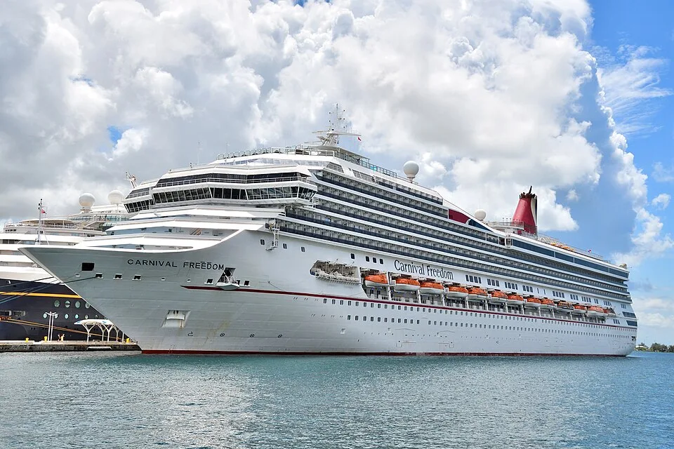 A large white cruise ship named "Carnival Freedom" floating on calm, turquoise water. The multi-deck vessel features a prominent bow, rows of balconies, orange lifeboats along the side, and a red funnel, with another ship partially visible behind it under a cloudy blue sky.