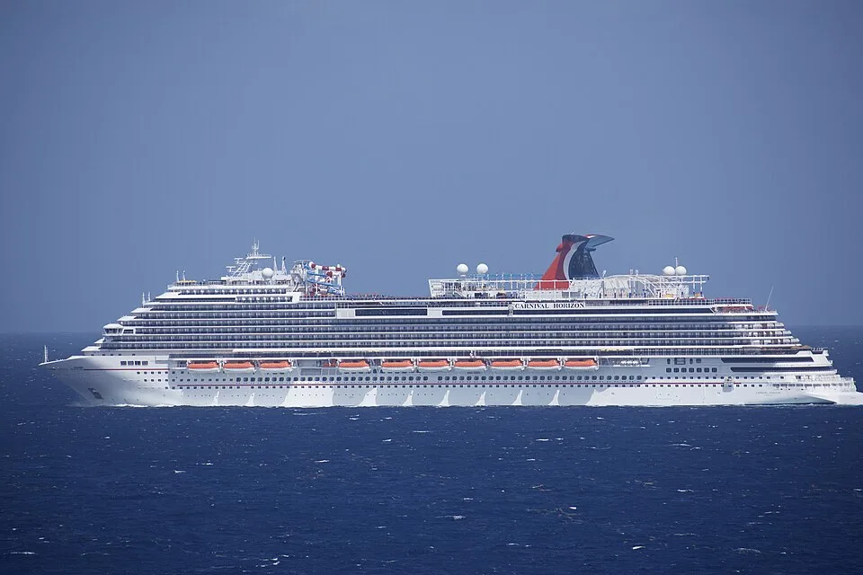A large white cruise ship named "Carnival Horizon" sailing across a deep blue ocean. The multi-deck ship features rows of orange lifeboats, a colorful water slide park on the top deck, and a signature red, white, and blue whale-tail funnel under a clear sky.