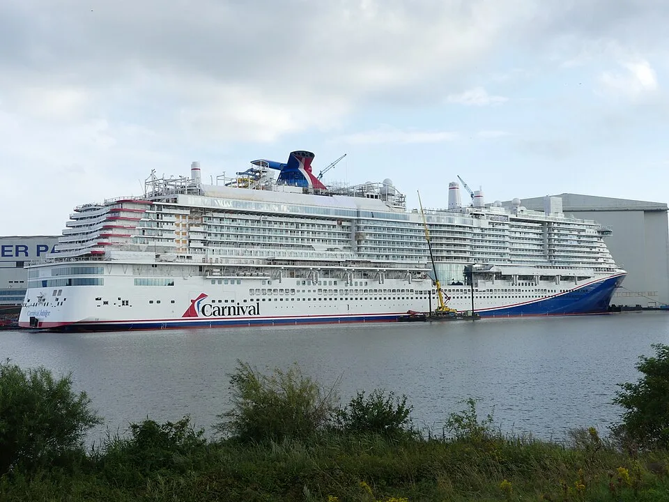 A massive white and blue Carnival cruise ship is docked in a shipyard or harbor under a cloudy sky. A large industrial building stands in the background, and green grassy bushes line the water in the foreground.