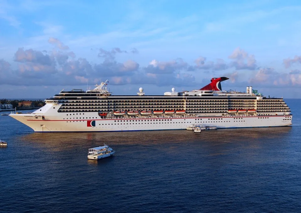 The large Carnival Legend cruise ship floats on calm blue water under a partly cloudy sky. The white multi-deck vessel features rows of small balconies, bright orange lifeboats along the side, its name near the bow, and a signature red and blue whale-tail funnel on top.