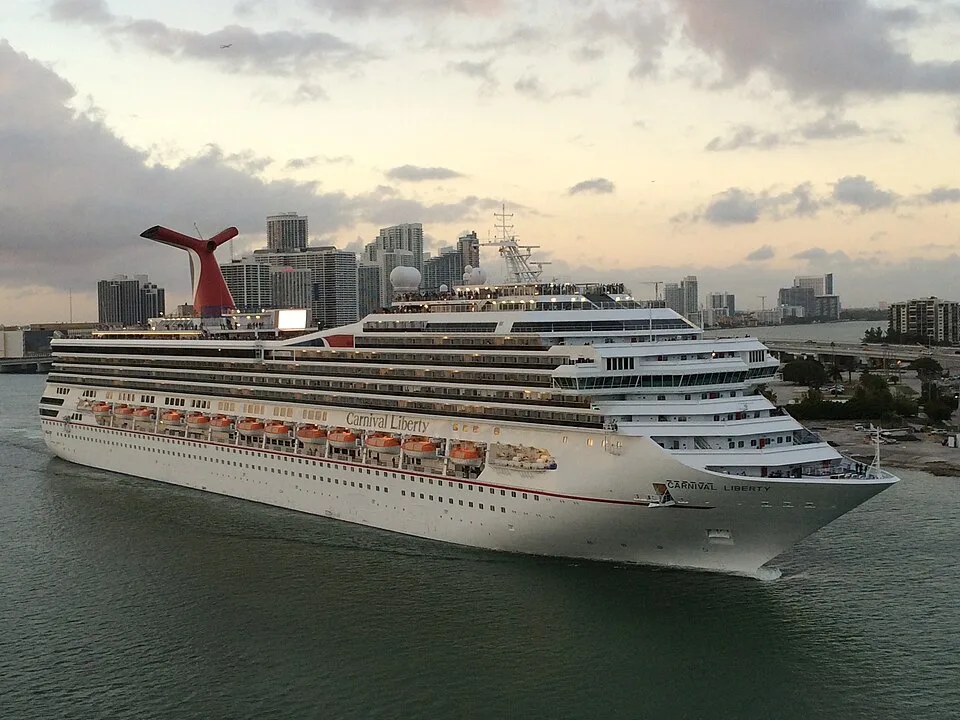 A large white cruise ship named "Carnival Liberty" sailing on calm water near a city. The massive ship features many tiered decks with orange lifeboats along the side, and a signature red whale-tail smokestack on top, with skyscrapers visible in the background under a cloudy sunset sky.