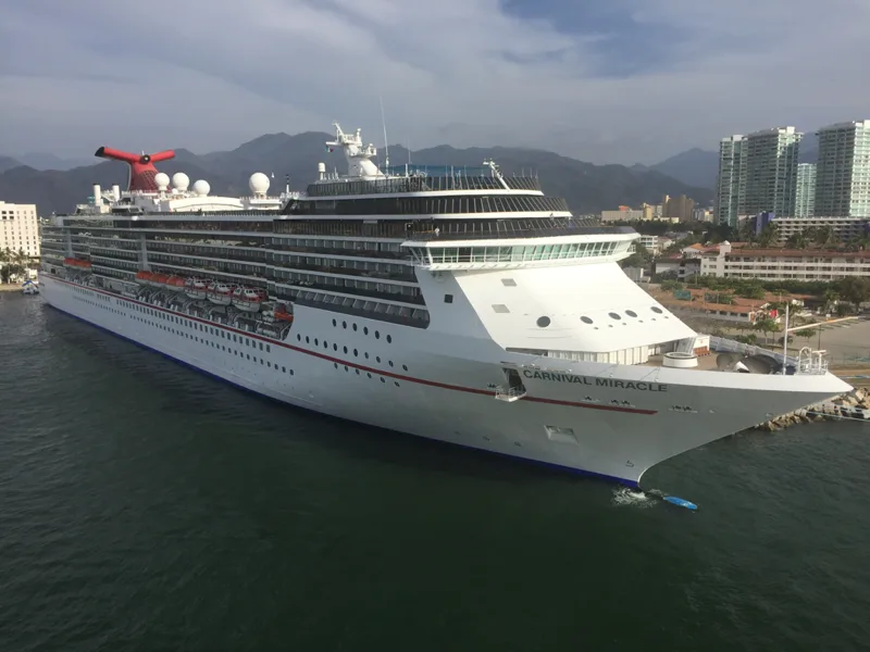 A large white cruise ship named "Carnival Miracle" docked in a harbor. The multi-deck vessel features a pointed bow, orange lifeboats along the side, and a signature red whale-tail smokestack, with coastal high-rises and mountains under a cloudy sky in the background.