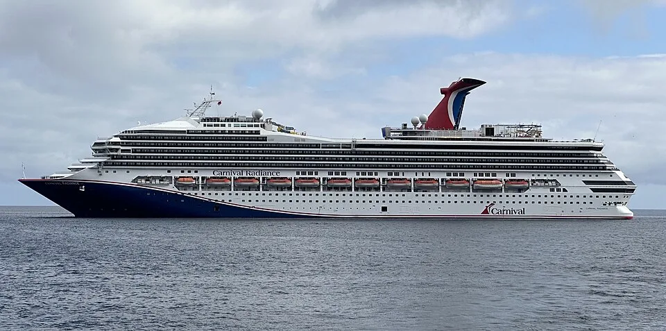 The large Carnival Radiance cruise ship sails on calm, open water under a overcast, cloudy sky. The vessel features a blue hull at the front, a white body with multiple rows of cabin windows, orange lifeboats, and its signature red, white, and blue funnel.
