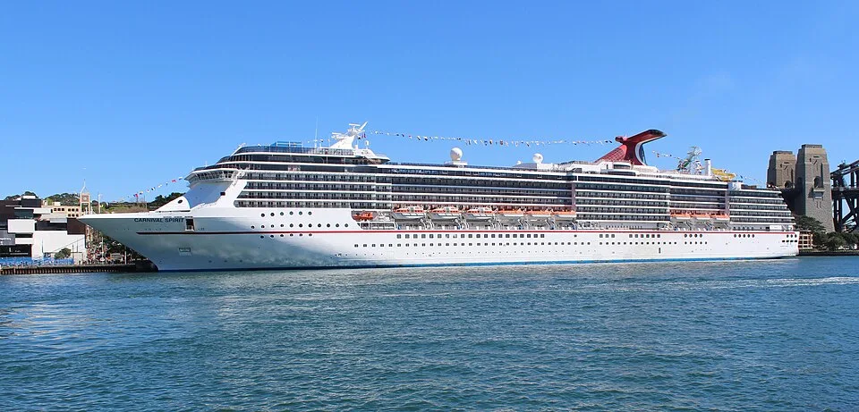 The large Carnival Spirit cruise ship sits docked in blue water under a clear sky. The white multi-deck vessel features rows of balconies, orange lifeboats along its side, a festive string of flags, and a signature red and blue whale-tail funnel near the rear.
