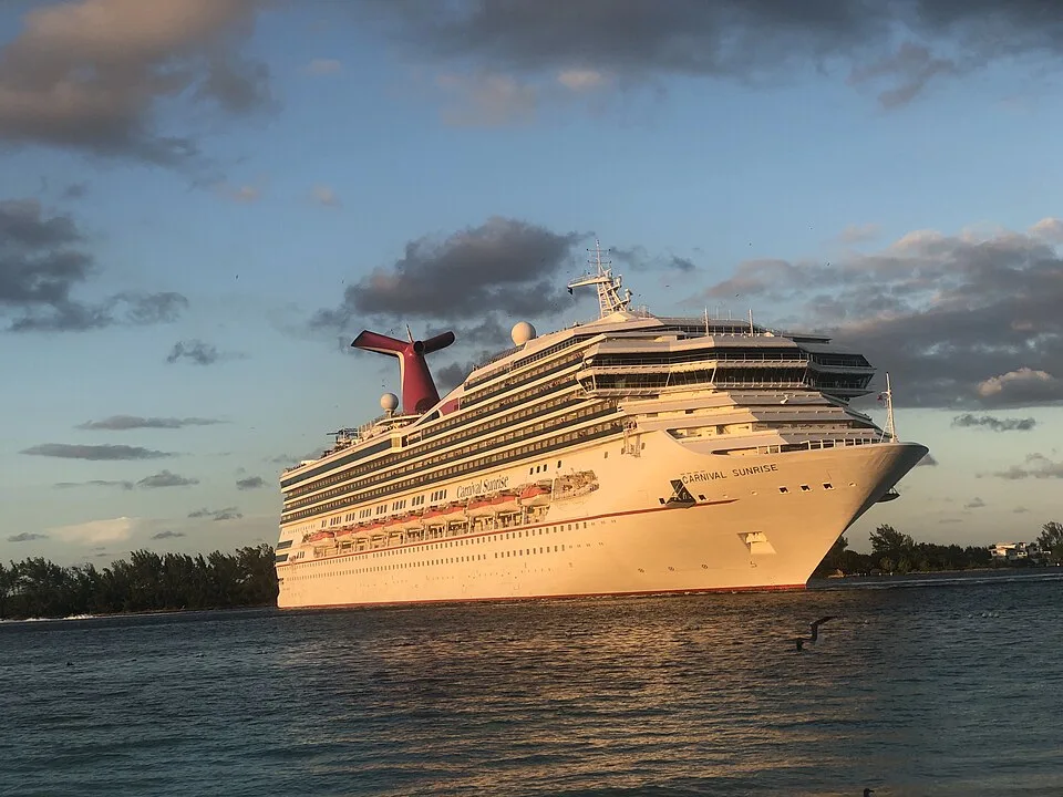 A large white cruise ship named "Carnival Sunrise" navigating calm water near a treelined shoreline. The multi-deck vessel features a prominent bow, orange lifeboats along the side, and a red whale-tail smokestack on top, captured under a partly cloudy sky during a late afternoon.