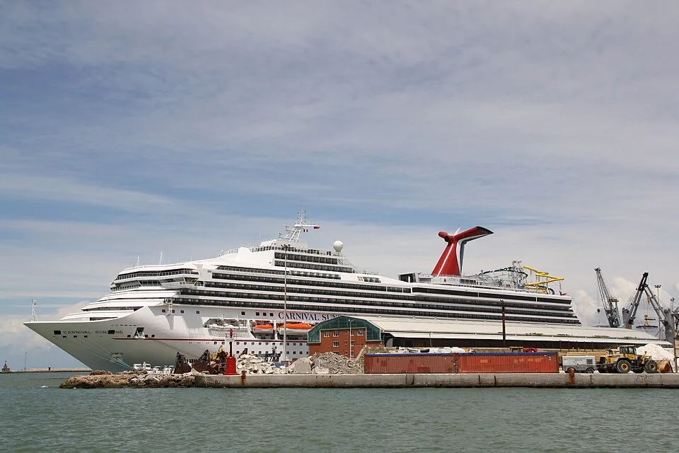 The large white Carnival Sunshine cruise ship is docked at an industrial port next to shipping containers and construction equipment. The ship features its signature red, white, and blue whale-tail funnel and multiple passenger decks under a partly cloudy sky.