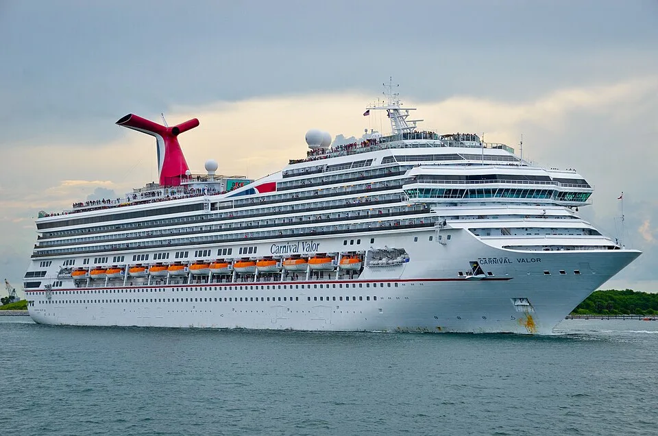 The massive Carnival Valor cruise ship sails through calm green water under a cloudy sky. The white multi-deck vessel features orange lifeboats along its side, the ship's name near the bow, crowds of people on the upper decks, and a prominent red and blue funnel.