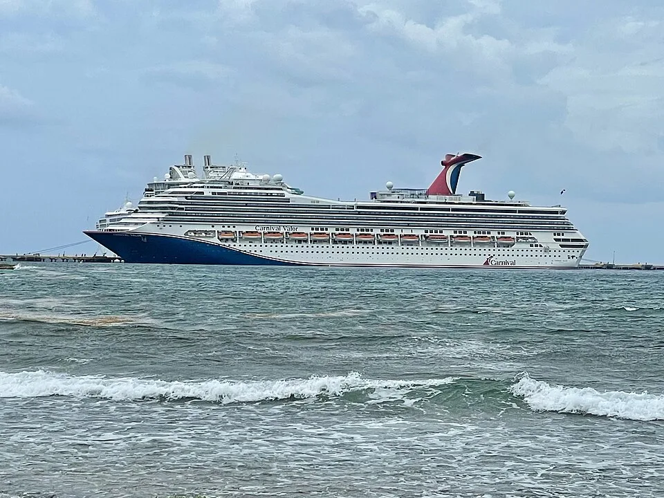 The massive Carnival Valor cruise ship sails on calm blue water under a cloudy sky. The vessel features a dark blue bow, multiple stacked tiers of cabin balconies, orange lifeboats along the side, and Carnival's signature red, white, and blue whale-tail funnel at the rear.