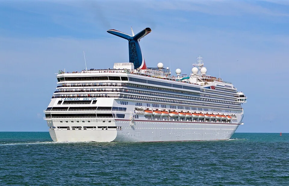 The Carnival Valor cruise ship sails through choppy blue-green water under a bright sky. The massive white multi-deck vessel features rows of balconies, orange lifeboats along the side, the ship's name visible on the stern, and a prominent blue and red whale-tail funnel on top.