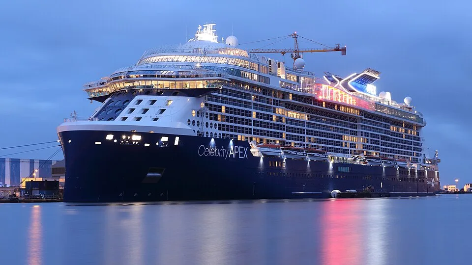 The Celebrity Apex cruise ship lit up at dusk, docked in calm water with its lights reflecting on the surface. The large, modern ship features a dark blue hull and multiple glowing decks against a twilight sky.