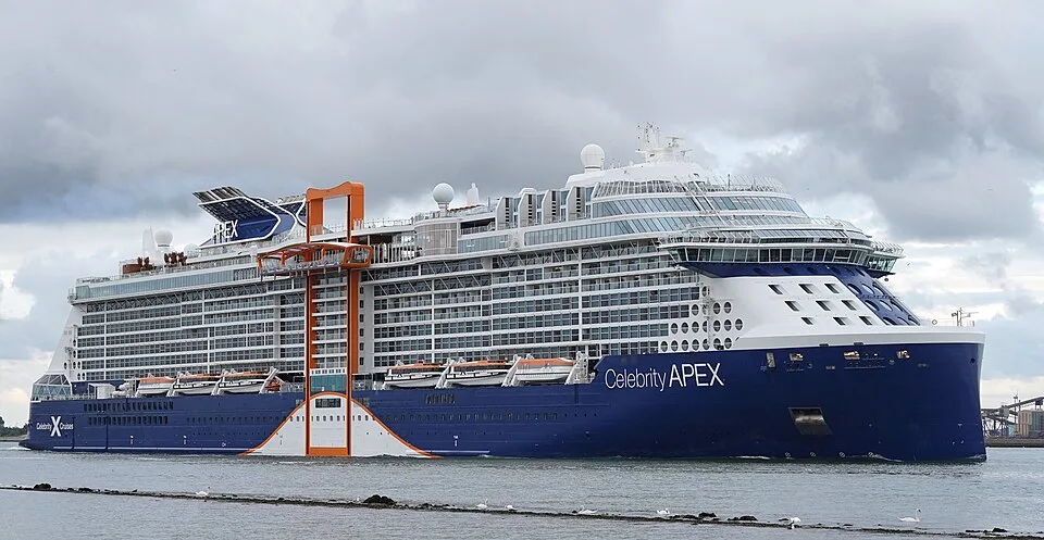 The massive Celebrity Apex cruise ship sails on calm water under a cloudy sky. The vessel features a striking dark blue hull, multiple decks of glass balconies, a unique orange cantilevered platform on its side, and its name clearly written near the bow.