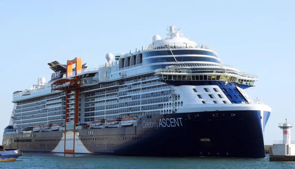 The massive modern cruise ship Celebrity Ascent sails on blue water under a clear sky. The ship features a dark blue and white hull, multiple stacked decks with tiered balconies, a prominent orange vertical structure on the side, and a small white lighthouse visible on the right.