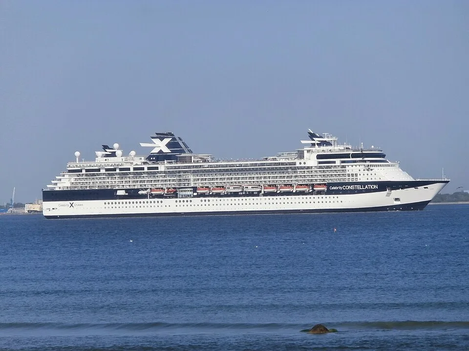The Celebrity Constellation cruise ship sailing on bright blue water under a clear sky. The large white and dark blue vessel features multiple decks, a line of orange lifeboats, and a distinctive "X" logo on its funnel.