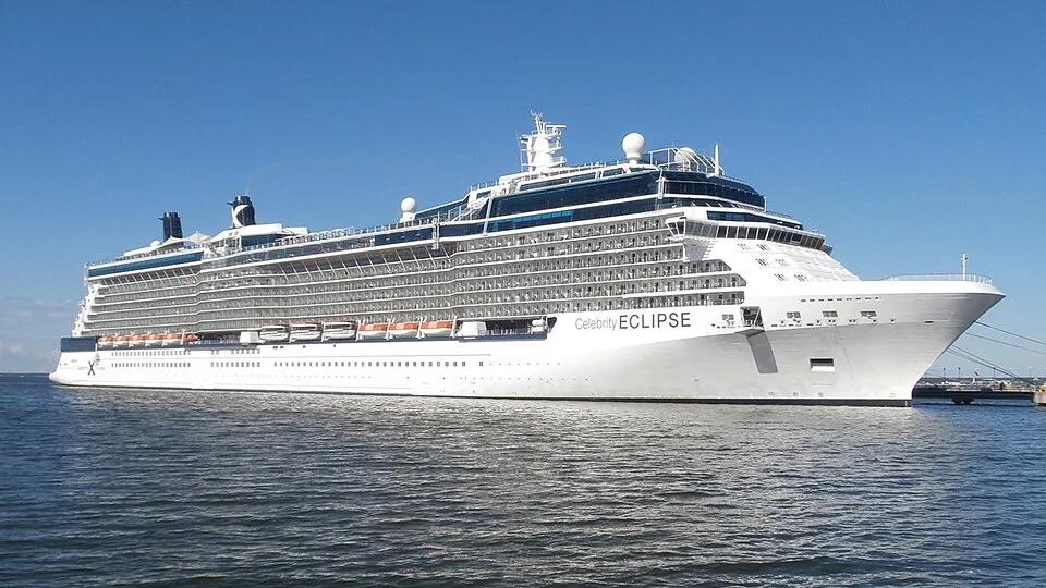 The massive white and blue cruise ship Celebrity Eclipse is docked in calm water next to a pier. The ship features multiple rows of passenger balconies, orange lifeboats along the lower side, and two dark smokestacks near the rear under a clear blue sky.