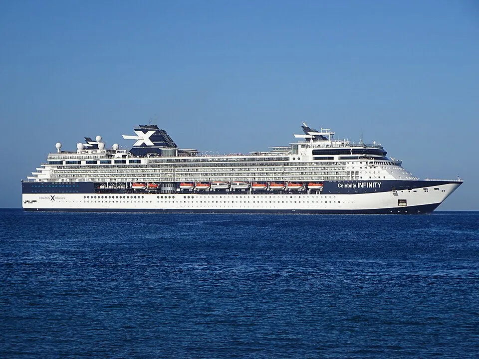 The large Celebrity Infinity cruise ship glides across deep blue water under a clear, bright sky. The vessel features a dark blue and white hull, rows of balconies, orange lifeboats along the side, and a dark funnel near the rear marked with a large white "X".