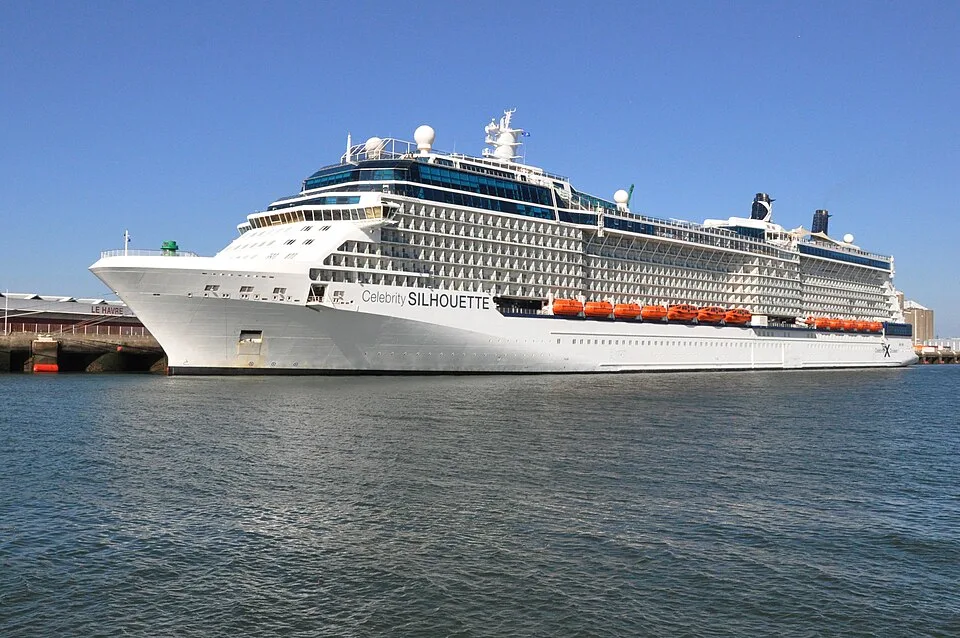 A large white cruise ship named "Celebrity SILHOUETTE" is docked on calm blue water under a clear blue sky. The multi-level ship features rows of windows, balconies, orange lifeboats along the side, and a dark funnel near the back. The bow points to the left near a distant harbor building.