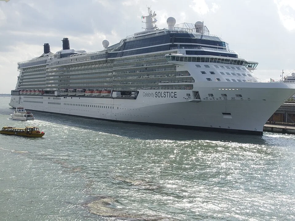 The Celebrity Solstice cruise ship docked in a harbor with shimmering water in the foreground. Two smaller tour boats navigate nearby. The massive white cruise vessel features multiple balcony decks and dark glass modern design accents.