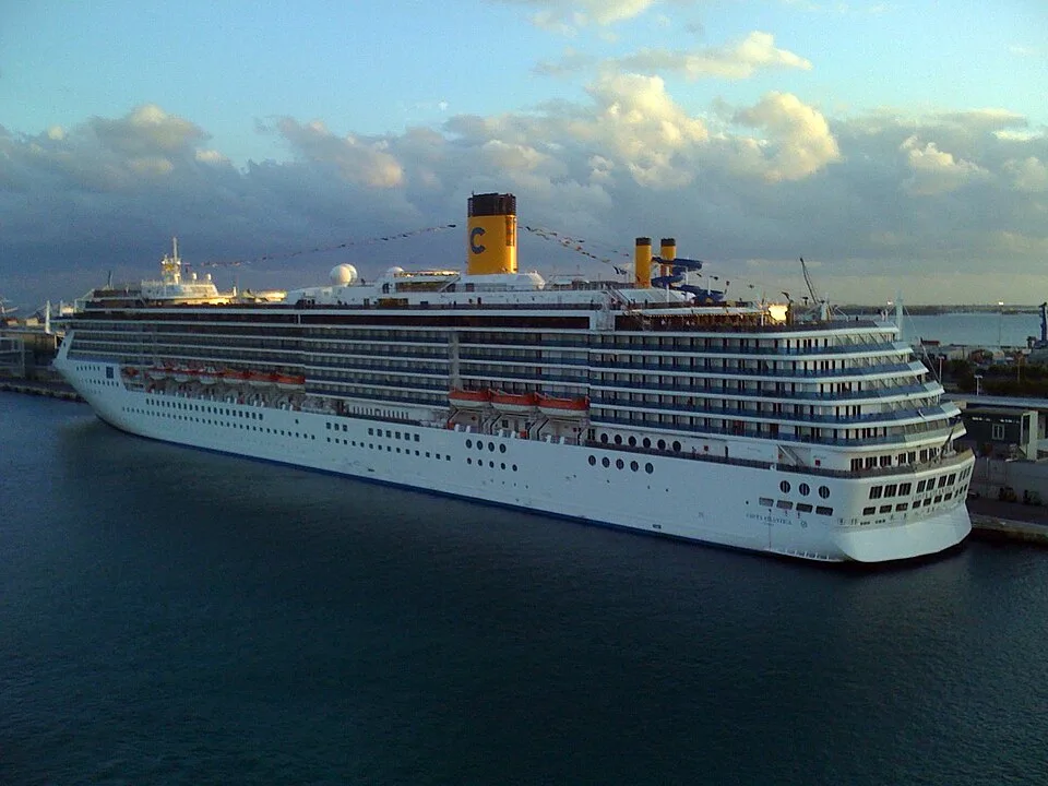 A large white Costa cruise ship with a prominent yellow funnel marked with a blue 'C' is docked in a calm harbor. The ship features multiple rows of balconies and windows under a partly cloudy sky during late afternoon or early morning light.