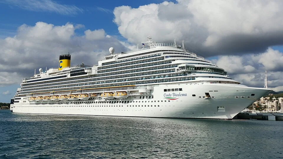 The massive white cruise ship Costa Diadema is docked in a port on calm blue water. The ship features numerous rows of glass passenger balconies, yellow lifeboats along its side, a yellow funnel with a blue "C" logo, and a background of fluffy white clouds in a blue sky.