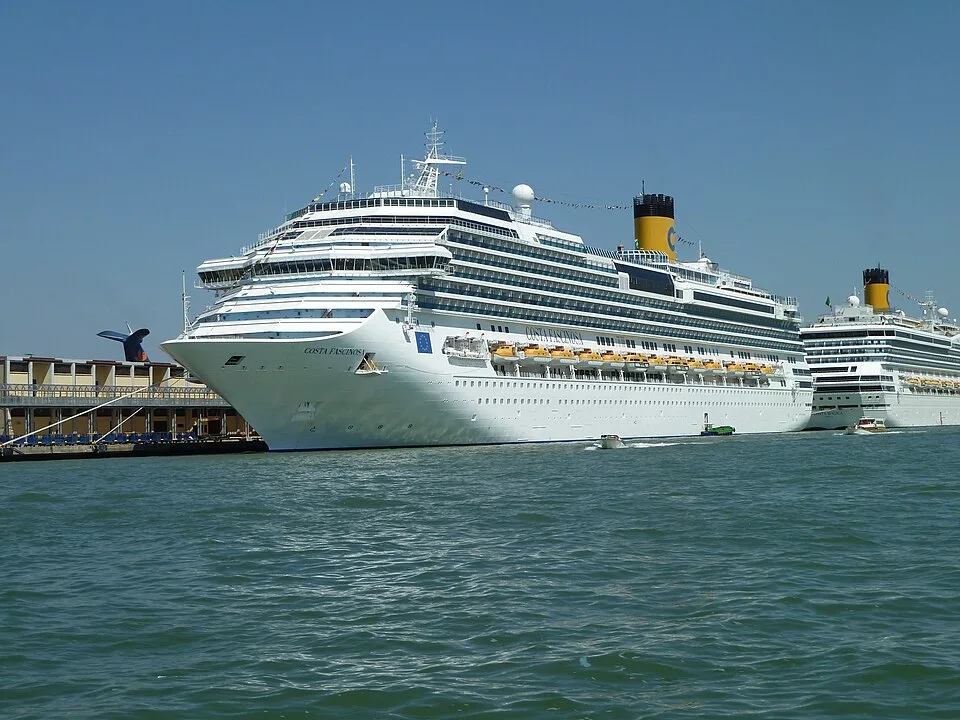 A large white cruise ship named "COSTA FASCINOSA" is docked in the water under a clear blue sky. It has many decks, rows of balconies, yellow lifeboats, and a tall yellow funnel. Part of a harbor building is on the left, and another Costa ship is visible further back to the right.