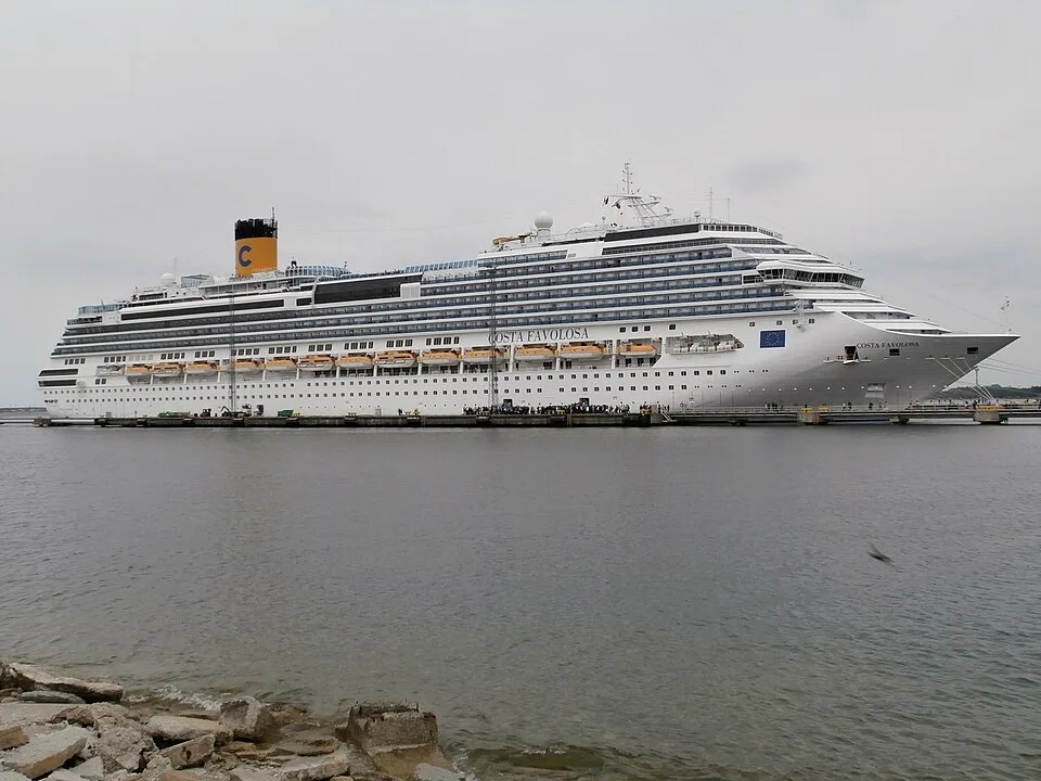 The Costa Favolosa cruise ship docked alongside a long pier. The large white vessel features numerous passenger decks, a row of yellow lifeboats, and a bright yellow funnel with a blue letter "C" on top, under an overcast sky.