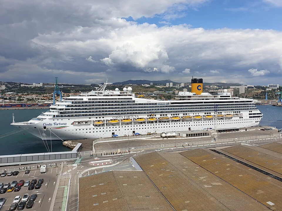 The large white cruise ship Costa Fortuna is docked at a city port terminal. The ship features its name on the side, yellow lifeboats, and a bright yellow funnel with a blue "C" logo. A parking lot and warehouse roofs sit in the foreground under a cloudy sky.