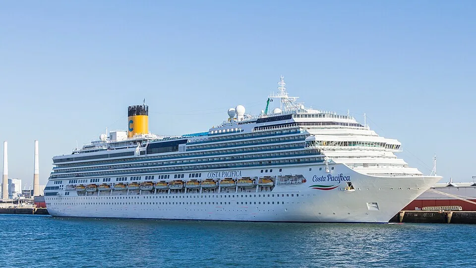 A large white cruise ship named "Costa Pacifica" sits on blue water under a clear sky. The multi-deck ship features rows of small windows, yellow lifeboats along its side, and a large yellow smokestack with a blue "C" logo near the back. A pier and industrial towers are visible in the background.