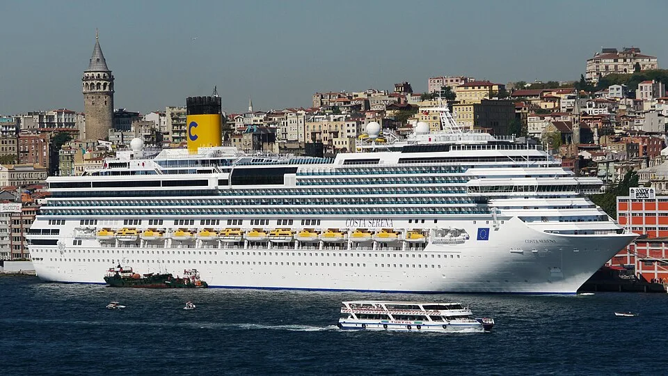 The Costa Serena cruise ship sailing past a coastal city. The large white ship features a yellow funnel with a blue "C" and yellow lifeboats. A smaller tour boat cruises in front of it, and a tall stone tower rises from the city hillside.